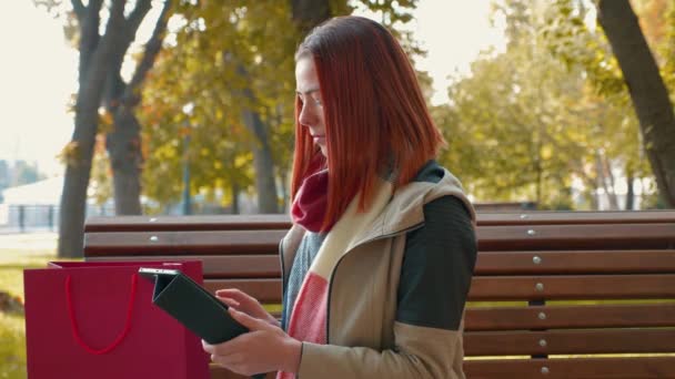 公园里穿着夹克的年轻迷人的红头发女人正在网上购物 在阳光灿烂的天气里 坐在长椅上的小女孩用平板电脑在网上下订单 零售业的概念 — 图库视频影像