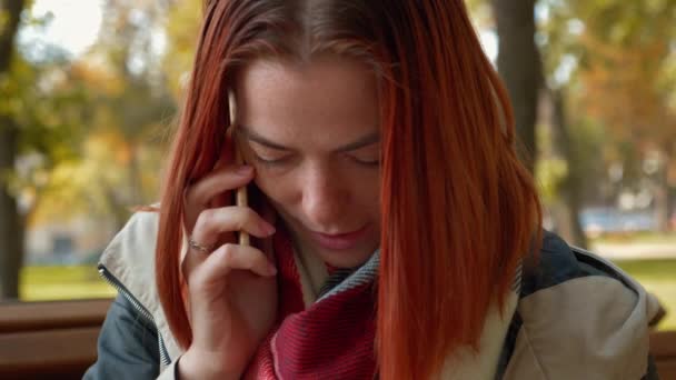 在停车场的红头发女人在用智能手机说话 戴狐狸毛的女孩坐在长椅上 在电话里和一家网上商店的经理交谈 在网上下订单 零售业的概念 — 图库视频影像