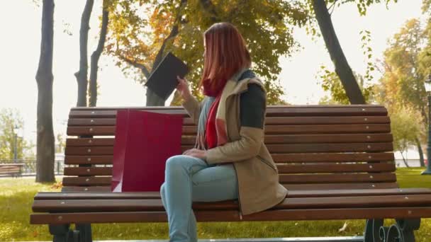 公園で若い魅力的な赤い髪の女性はオンラインショッピングです 偽の髪の女の子がベンチに座って 赤い紙袋からタブレットを取り出し それを使用し始めます 小売業の概念 4K映像 — ストック動画