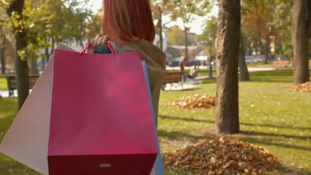穿着夹克的年轻漂亮的红头发女人正在购物 一个留着狐狸毛的女孩带着各种颜色的纸袋穿过公园 零售业的概念 4K镜头 — 图库视频影像
