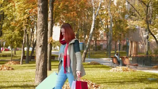 穿着夹克的年轻漂亮的红头发女人正在购物 一个留着狐狸毛的女孩带着各种颜色的纸袋穿过公园 零售业的概念 4K镜头 — 图库视频影像