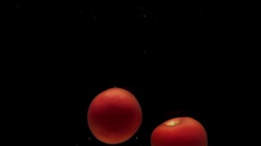 İki kırmızı domates siyah zemin üzerinde şeffaf suya düşüyor. Akvaryuma taze organik sebzeler sıçrıyor. Market, sağlıklı yiyecekler, diyet, vejetaryenlik, hava kabarcıkları. Ağır çekim. Yakın plan.