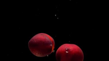 İki olgun şeftali, siyah arka planda şeffaf suya dökülüyor. Akvaryuma taze organik meyveler sıçrıyor. Market, sağlıklı yiyecekler, diyet, vejetaryenlik, hava kabarcıkları. Ağır çekim. Yakın plan..