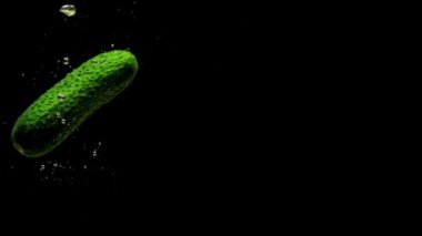 Yeşil salatalık siyah arkaplanda saydam suya düşüyor. Akvaryuma taze organik sebzeler sıçrıyor. Market, sağlıklı yiyecekler, diyet, vejetaryenlik, hava kabarcıkları. Ağır çekim. Yakın plan..