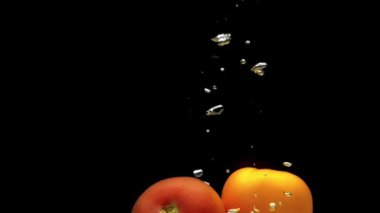 Kırmızı ve sarı domatesler siyah arkaplanda saydam suya düşüyorlar. Akvaryuma taze organik sebzeler sıçrıyor. Market, sağlıklı yiyecekler, vejetaryenlik, hava kabarcıkları. Ağır çekim. Yakın plan..