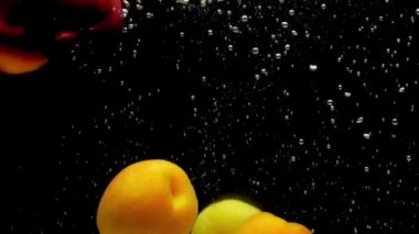 Şeftali ve kayısılar siyah arka planda şeffaf suya düşüyorlar. Akvaryuma taze organik meyveler sıçrıyor. Market, sağlıklı yiyecekler, diyet, vejetaryenlik, hava kabarcıkları. Ağır çekim. Yakın plan..