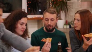 Happy friends talking, eating pizza, drinks alcohol and celebrating new years eve party at home. Bottles of beer and glasses of mulled wine on the table. Holidays and celebration concept. 4K footage.