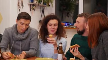 Happy friends talking, eating pizza, drinks alcohol and celebrating new years eve party at home. Bottles of beer and glasses of mulled wine on the table. Holidays and celebration concept. 4K footage.
