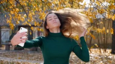 Genç çekici esmer kadın, sonbaharda sarı ağaçların arka planında akıllı telefondan selfie çekiyor. Güneşli bir günde yeşil kazaklı güzel bir kız parkın ortasında fotoğraflandı. 4k görüntü