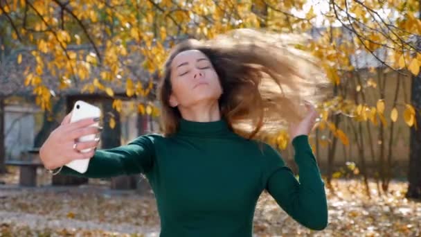 一个年轻迷人的黑发女人在黄树的背景下 在智能手机上自拍 阳光灿烂的一天 一位穿着绿色毛衣的漂亮姑娘在公园中央拍照 4K镜头 — 图库视频影像