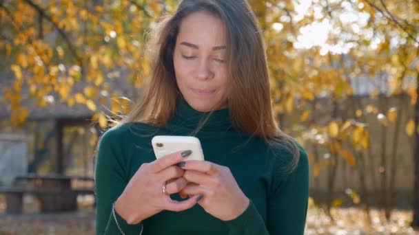Молодая Привлекательная Брюнетка Использует Белый Смартфон Фоне Пожелтевших Деревьев Осенью — стоковое видео