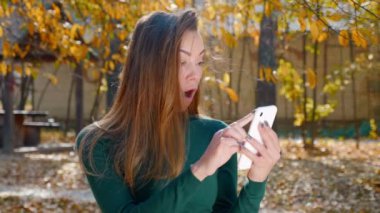 Genç çekici esmer kadın akıllı telefonda gördüklerine şaşırdı. Sonbaharda güneşli bir günde yeşil kazaklı güzel bir kız parkın ortasında indirimleri izliyor. 4k