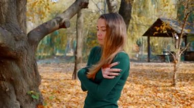 Donmuş genç ve çekici esmer kadın sonbaharda sarı ağaçların arka planında parkta yürüyor. Yeşil kazaklı güzel bir kız güneşli bir günde ormanın ortasında geziniyor. 4k