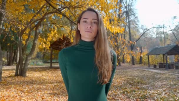 秋には黄色の木々の背景に若い魅力的なブルネットの女性の肖像画 緑のセーターを着た可愛い女の子が晴れた日に森の真ん中に立っている 4K映像 — ストック動画