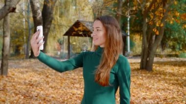 Genç ve çekici esmer kadın akıllı telefonundan sosyal medyaya video yayınlıyor. Yeşil kazaklı güzel bir kız sonbaharda güneşli bir günde parkın ortasında bir video görüşmesi yapar. 4k
