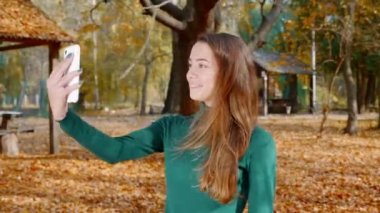 Genç ve çekici esmer kadın akıllı telefonundan sosyal medyaya video yayınlıyor. Yeşil kazaklı güzel bir kız sonbaharda güneşli bir günde parkın ortasında bir video görüşmesi yapar. 4k
