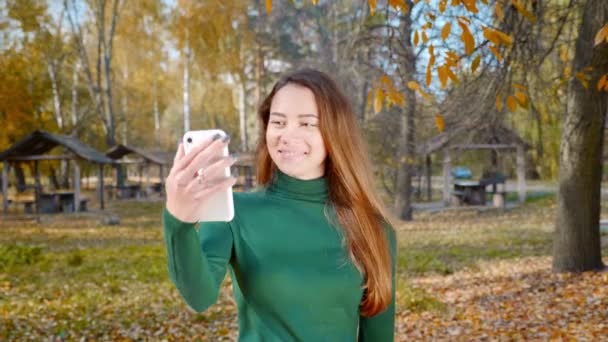 年轻迷人的黑发女人走路 并从她的智能手机向社交媒体传输视频 秋天的一个阳光灿烂的日子 身穿绿色毛衣的漂亮姑娘在公园中央打了一个视频电话 — 图库视频影像