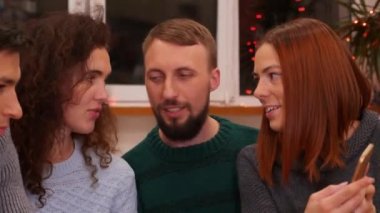 Arkadaşlarına fotoğraflarını gösteren hanımefendinin yakın plan fotoğrafları, akıllı telefondan çekilmiş videoları. Mutlu arkadaşlar telefonla izliyor, şenlik masasında konuşuyor ve evde yeni yıl partisini kutluyorlar. Tatil konsepti. 4k