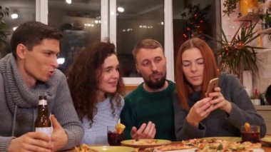 Genç bayan arkadaşlarına resimlerini, akıllı telefondaki videolarını gösteriyor. Mutlu arkadaşlar telefonla izliyor, şenlik masasında konuşuyor ve evde yeni yıl partisini kutluyorlar. Tatil konsepti 4k video