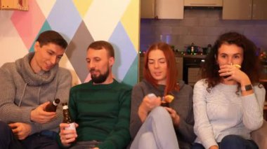 Mutlu arkadaşlar kanepede oturuyor, alkol içiyor ve evde yeni yıl partisini kutluyorlar. Çok renkli ışıklar, arka planda çelenkler. Tatiller ve kutlama konsepti. 4k görüntü.