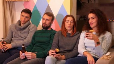 Mutlu arkadaşlar kanepede oturuyor, alkol içiyor ve evde yeni yıl partisini kutluyorlar. Çok renkli ışıklar, arka planda çelenkler. Tatiller ve kutlama konsepti. 4k görüntü.
