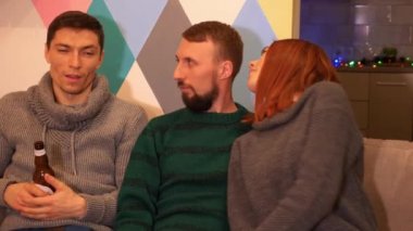 Mutlu arkadaşlar kanepede oturuyor, alkol içiyor ve evde yeni yıl partisini kutluyorlar. Çok renkli ışıklar, arka planda çelenkler. Tatiller ve kutlama konsepti. 4k görüntü.