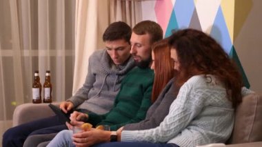 Mutlu arkadaşlar kanepede oturuyor, tablet kullanıyor ve evde yeni yıl partisini kutluyorlar. Çok renkli ışıklar, arka planda çelenkler. Tatiller ve kutlama konsepti. 4k görüntü.