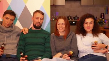 Mutlu arkadaşlar kanepede oturuyor, alkol içiyor ve evde yeni yıl partisini kutluyorlar. Çok renkli ışıklar, arka planda çelenkler. Tatiller ve kutlama konsepti. 4k görüntü.