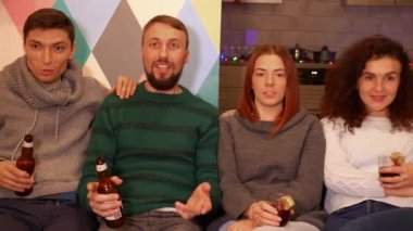 Mutlu arkadaşlar kanepede oturuyor, alkol içiyor ve yeni yıl tatillerinde TV 'de kış sporu maçı izliyorlar. Çok renkli ışıklar, arka planda çelenkler. Tatiller ve kutlama konsepti. 4k