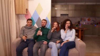 Mutlu arkadaşlar kanepede oturuyor, alkol içiyor ve yeni yıl tatillerinde televizyon izliyorlar. Çok renkli ışıklar, arka planda çelenkler. Tatiller ve kutlama konsepti. 4k görüntü.