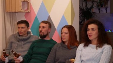 Mutlu arkadaşlar kanepede oturuyor, alkol içiyor ve yeni yıl tatillerinde televizyon izliyorlar. Çok renkli ışıklar, arka planda çelenkler. Tatiller ve kutlama konsepti. 4k görüntü.