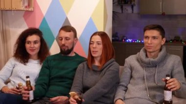 Mutlu arkadaşlar kanepede oturuyor, alkol içiyor ve yeni yıl tatillerinde televizyon izliyorlar. Çok renkli ışıklar, arka planda çelenkler. Tatiller ve kutlama konsepti. 4k görüntü.