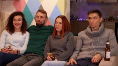 Mutlu arkadaşlar kanepede oturuyor, alkol içiyor ve yeni yıl tatillerinde televizyon izliyorlar. Çok renkli ışıklar, arka planda çelenkler. Tatiller ve kutlama konsepti. 4k görüntü.