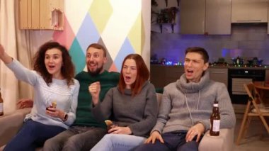 Mutlu arkadaşlar kanepede oturuyor, alkol içiyor ve yeni yıl tatillerinde TV 'de kış sporu maçı izliyorlar. Arka planda çok renkli duvar var. Tatiller ve kutlama konsepti. 4k görüntü.