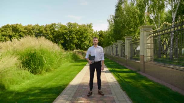 Επιχειρηματίας στέκεται σε ένα πάρκο με ένα φορητό υπολογιστή στα χέρια, κοιτάζοντας την κάμερα — Αρχείο Βίντεο