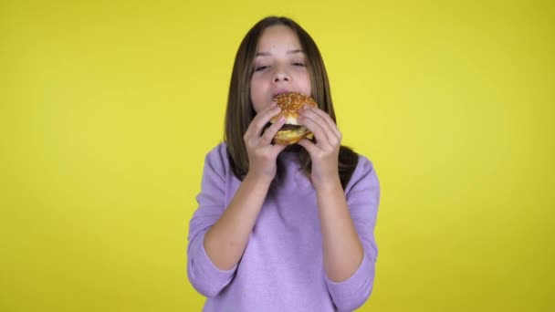 Nastolatka w różowym swetrze zjada hamburgera na żółtym tle z przestrzenią do kopiowania — Wideo stockowe