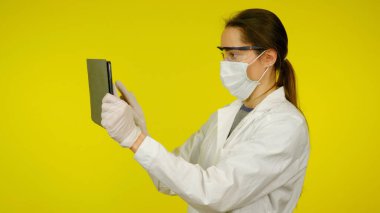 Tıbbi maskeli, gözlüklü ve lateks eldivenli genç doktor tablete bakıyor.
