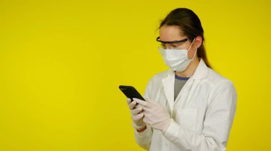Tıbbi maskeli, gözlüklü ve lateks eldivenli doktor akıllı telefona bakar.