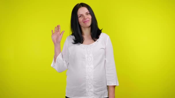 Olá ou adeus. Jovem grávida sorri e balança a mão sobre um fundo amarelo — Vídeo de Stock