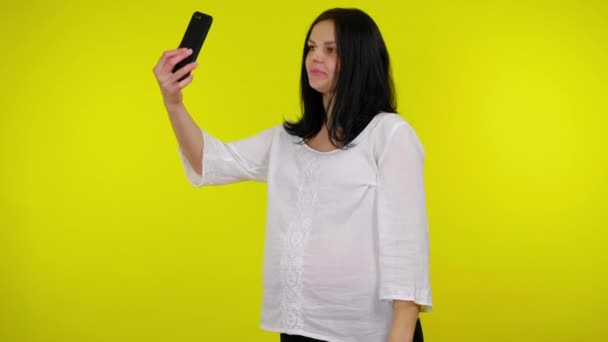 若い妊娠中の女性は黄色の背景にスマートフォンで自画撮り写真を撮る — ストック動画