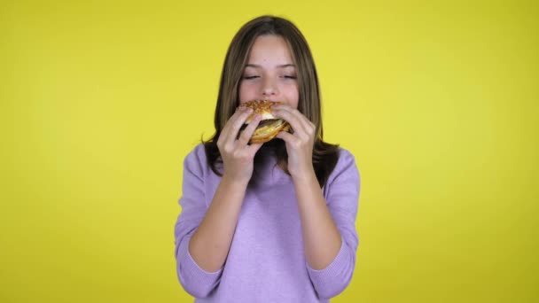 Tonåring flicka i en tröja äter en hamburgare och njuter av smaken på gul bakgrund — Stockvideo