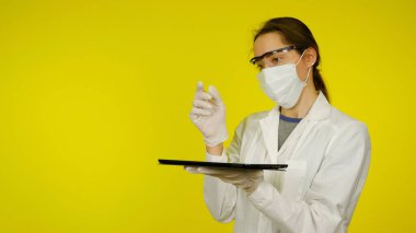 Tıbbi maskeli, gözlüklü ve lateks eldivenli doktor elinde bir tablet tutuyor.