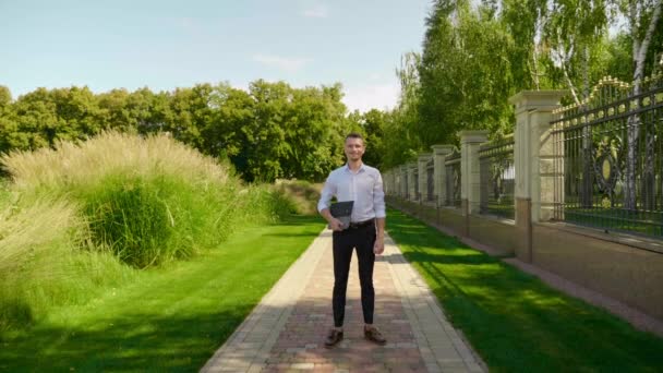 Uomo d'affari in un parco con un computer portatile in mano, guardando la fotocamera e sorridendo — Video Stock