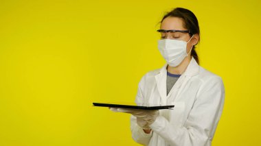 Tıbbi maskeli, gözlüklü ve lateks eldivenli doktor elinde bir tablet tutuyor.