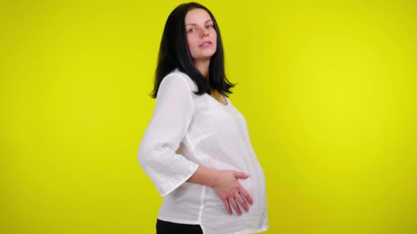 Молодая беременная женщина массирует живот руками на желтом фоне — стоковое видео