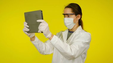 Tıbbi maskeli, gözlüklü ve lateks eldivenli genç doktor tablete bakıyor.