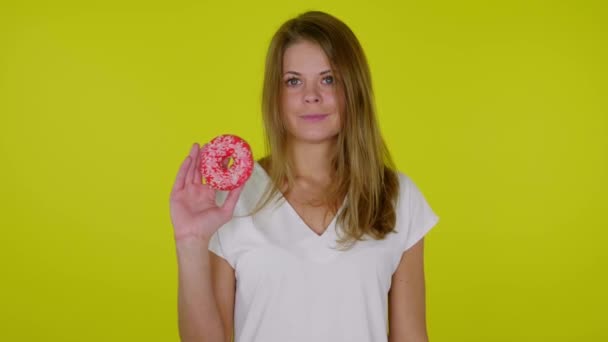 Mulher levanta a mão com um donuts vermelho, mostra como, sorri em um fundo amarelo — Vídeo de Stock