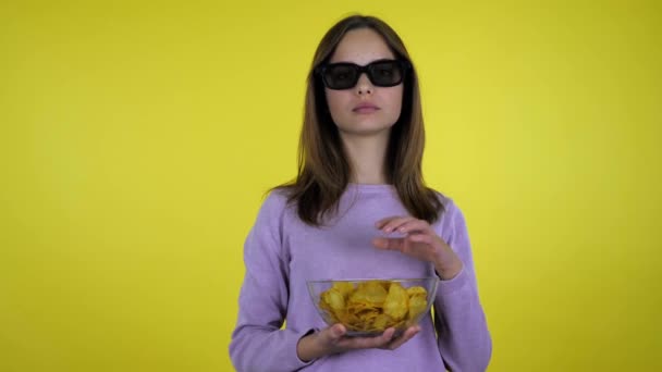 Meisje kijkt film in 3D bril en houdt chips in een hand in glazen schaal — Stockvideo