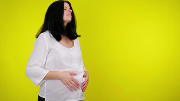 Молодая беременная женщина массирует живот с ребенком внутри на желтом фоне — стоковое видео