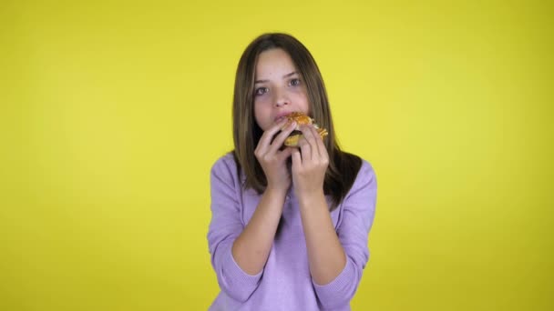 Tiener meisje in een roze trui beten en eet hamburger op gele achtergrond — Stockvideo